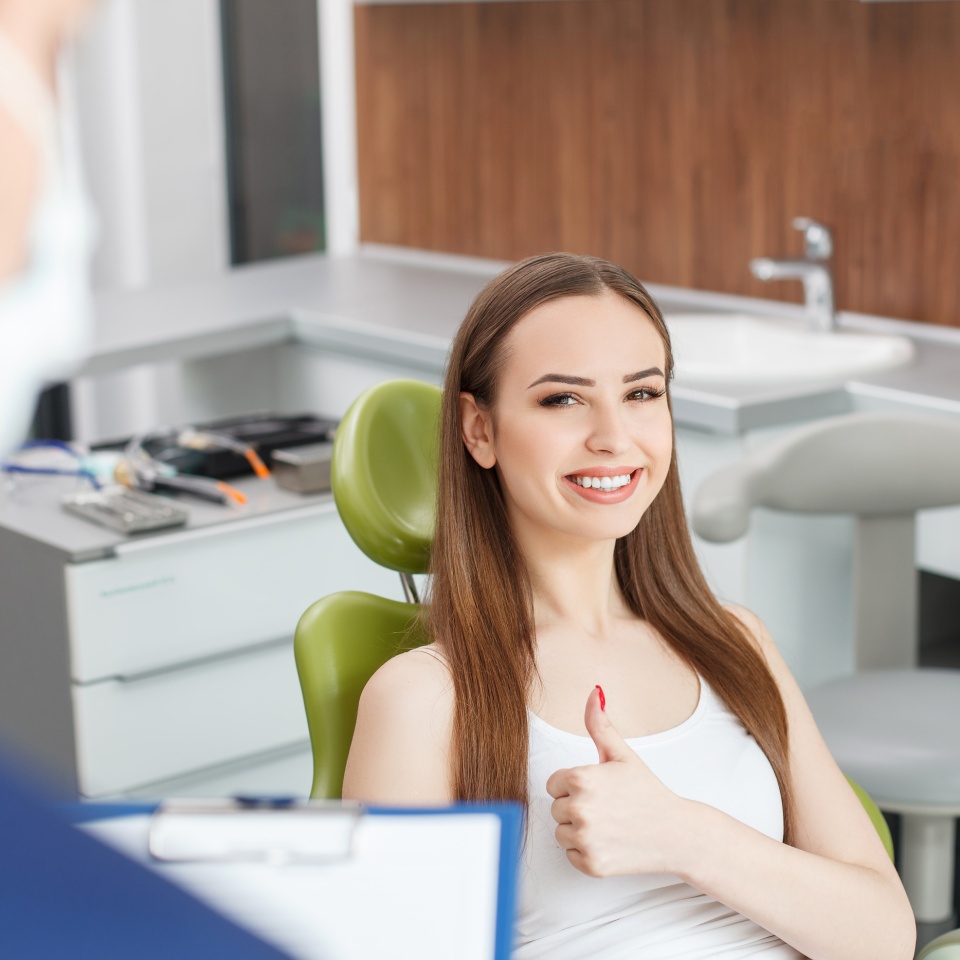 Beautiful healthy girl is sitting in chair and smiling. She is looking at her dentist and giving thumb up. The man is holding a folder and analyzing the results of treating