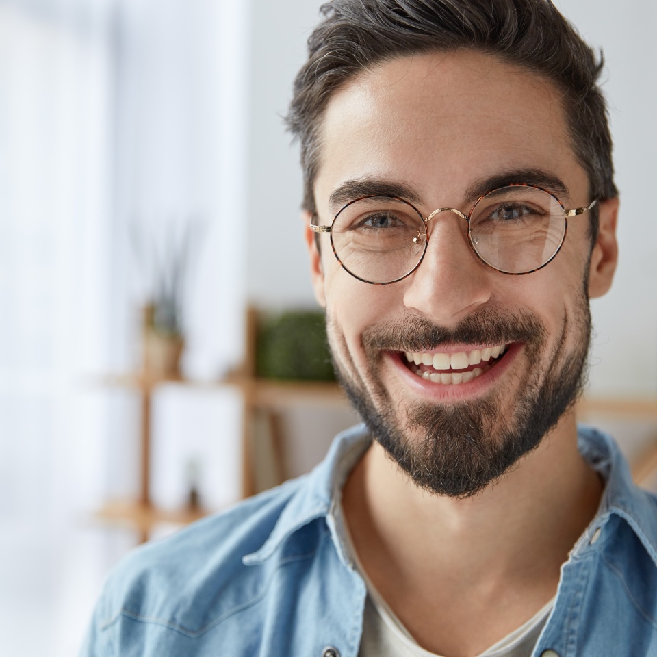 Close up shot of cheerful satisfied attractive male with stubble, has broad smile, wears round spectacles, rejoices success at work, stands against cozy interior. Fashionable designer glad be praised