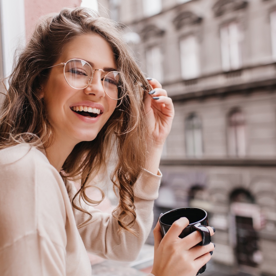 Close-up portrait of laughing brunette girl in beige sweater drinking coffee on city background. Lovable lady standing near window and looking at beautiful building with cup of tea.