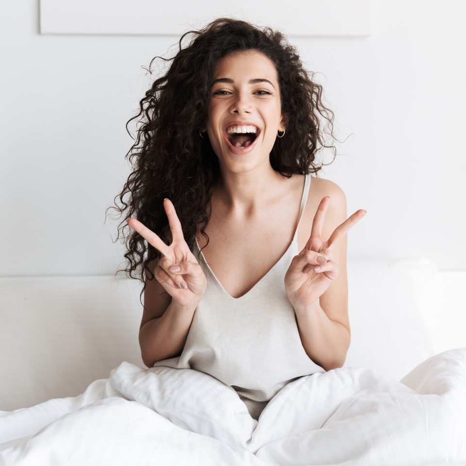 Happy young girl with dark curly hair sitting in bed in the morning and showing peace gesture