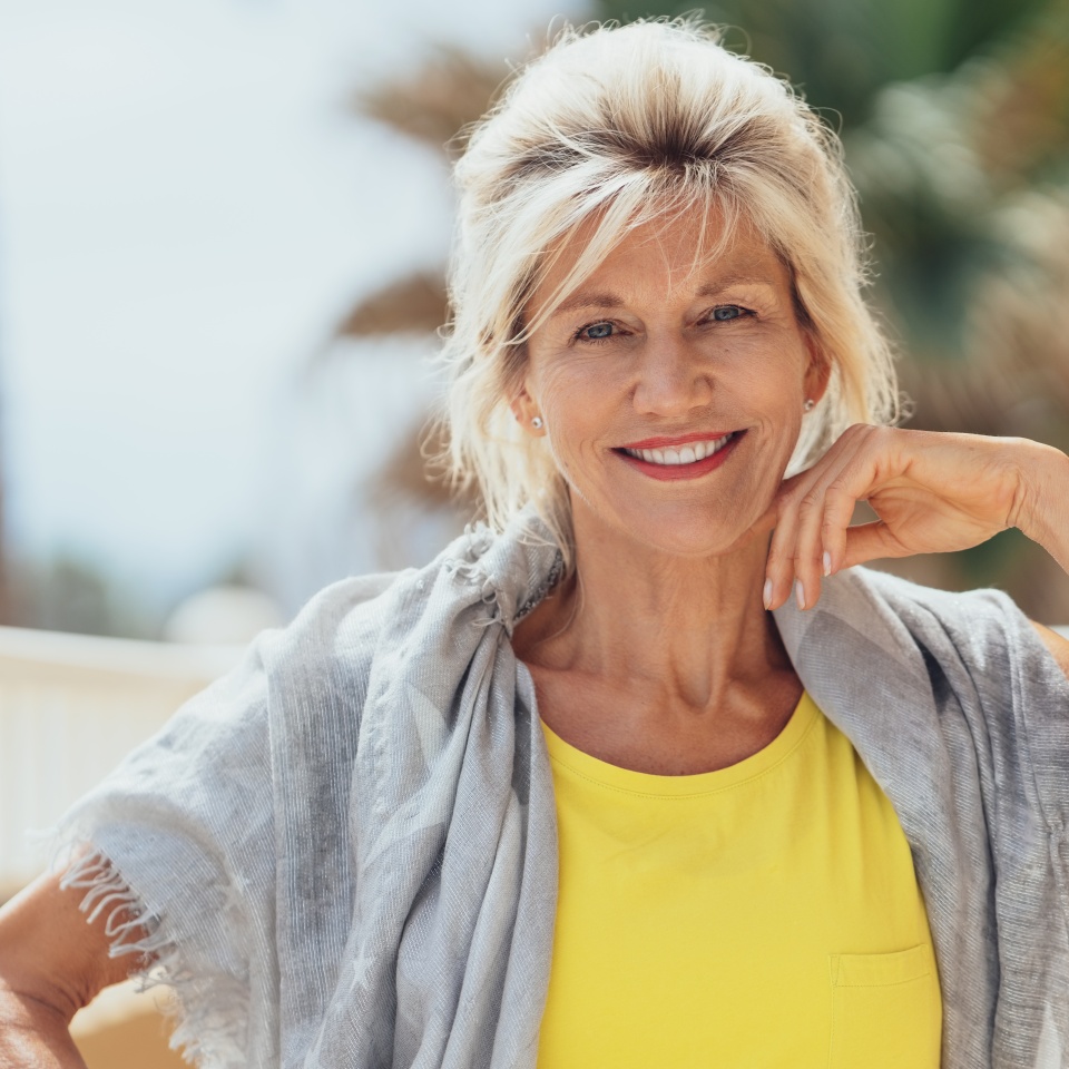 Stylish blond woman sitting on a balcony with a shawl over her shoulders in the sunshine smiling at the camera