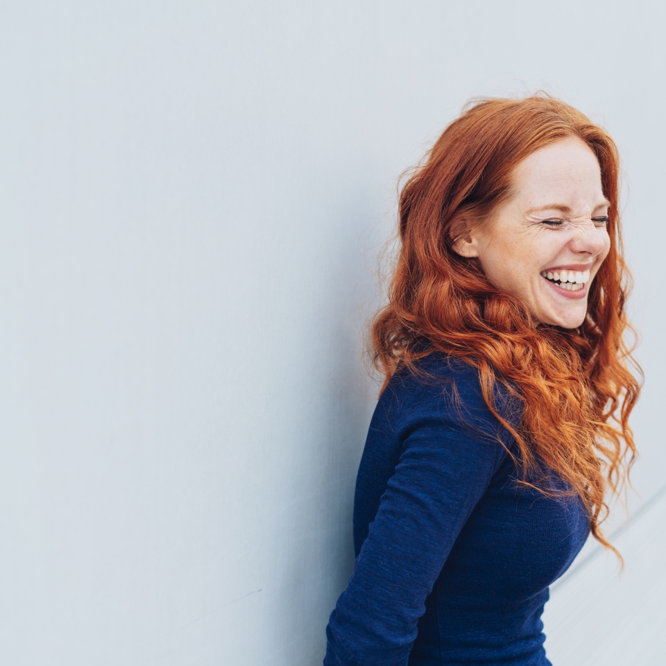 Attractive young woman standing giggling or laughing at something she finds very funny while leaning against a white exterior wall with copy space