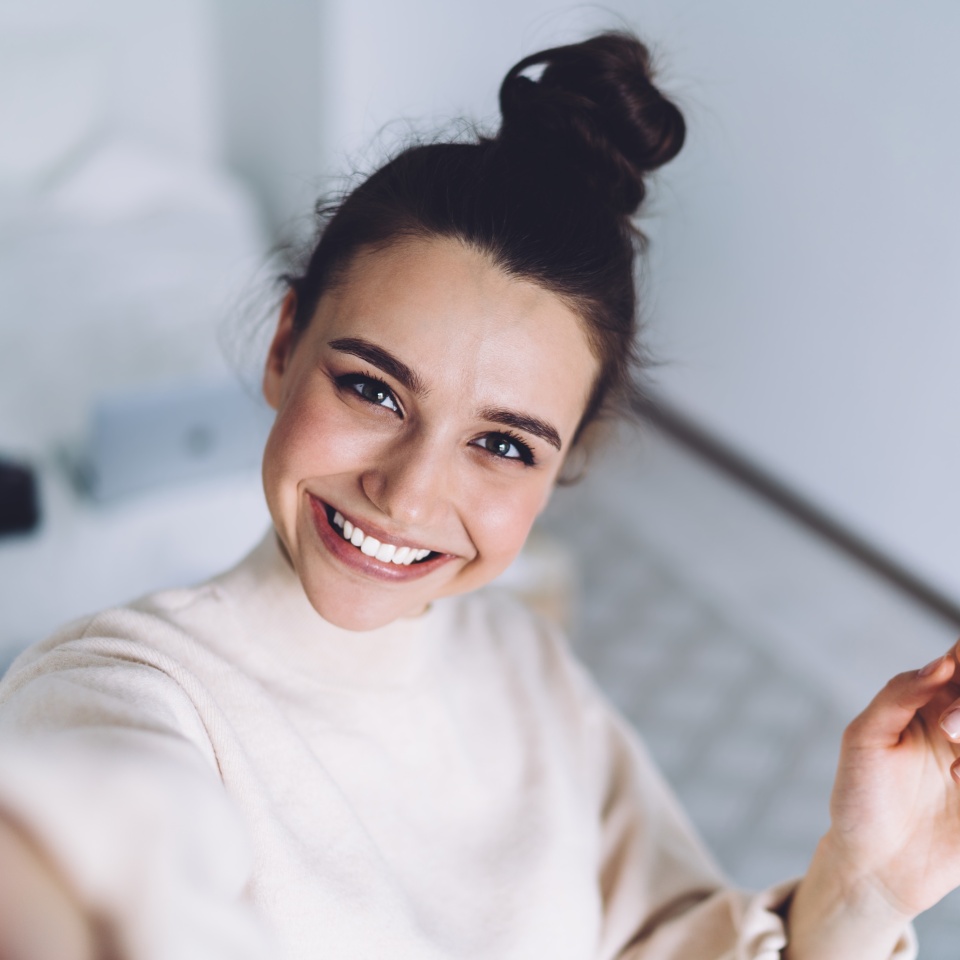 Pretty young female with big smile standing at bedroom after work with laptop and having fun taking light cheerful selfie on blurred background