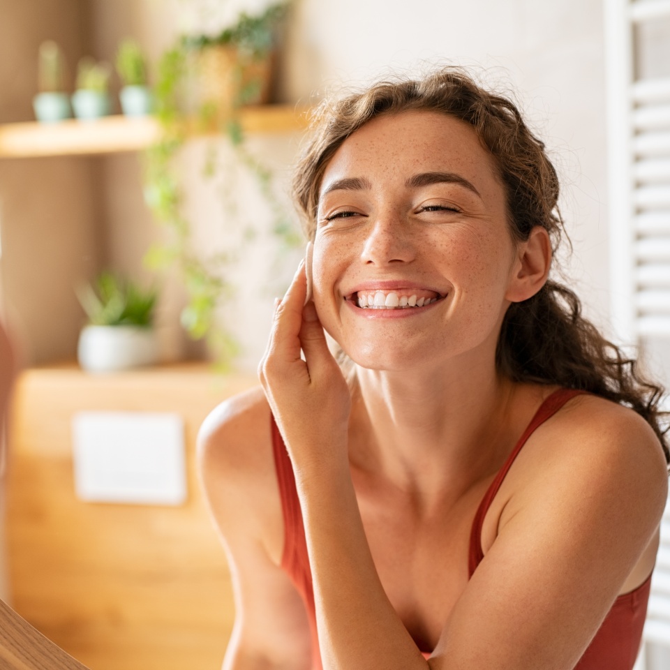 Happy smiling beautiful girl cleaning skin face with cotton pad. Beauty natural woman looking in mirror while cleansing skin face and using cosmetic products for properly deep clean.