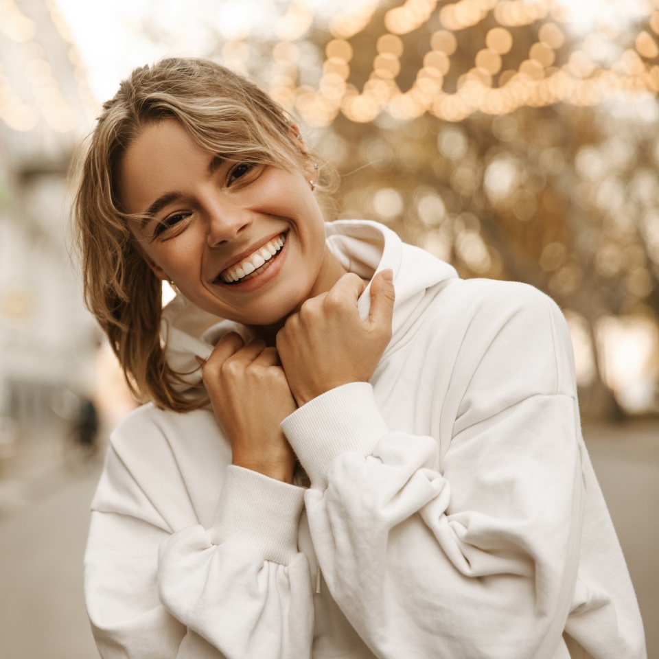 Close up portrait caucasian young happy woman with fresh and clean skin stands outside. Smiling blonde holds collar of white sweatshirt. Lifestyle, female beauty concept