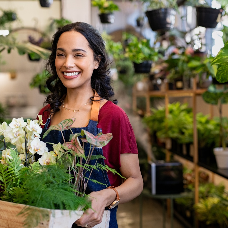Beautiful florist holding wooden box of fresh plants at flower shop. Latin hispanic woman working in floral shop with copy space. Successful florist smiling while holding fresh flowers and plants.