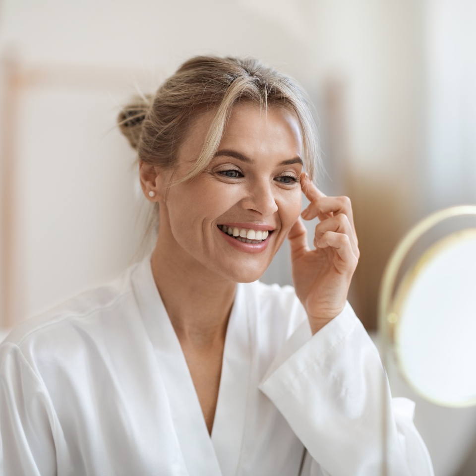 Cheerful beautiful blonde middle aged lady in silky bathrobe using moisturizing eye cream, looking at mirror and smiling, enjoying face care routine, closeup shot. Dark circles, eye wrinkles treatment