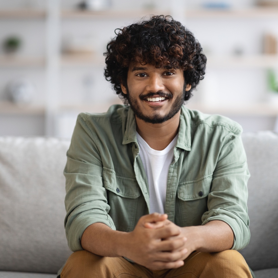 Cheerful millennial curly bearded indian guy in casual outfit sitting on couch at home, holding hands in lock and smiling at camera, young eastern man posing in living room, copy space