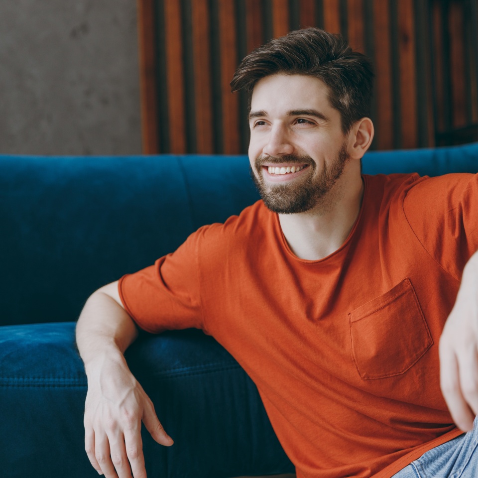 Young smiling pensive happy man wear red t-shirt look aside think sit on blue sofa couch stay at home hotel flat rest relax spend free spare time in living room indoors grey wall People lounge concept