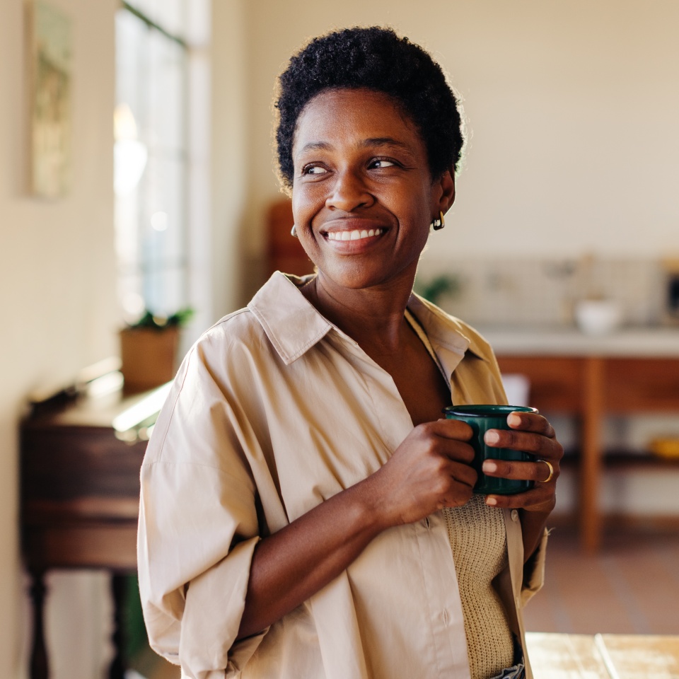Happy Brazilian woman looking outside the window in her home kitchen, smiling and holding an enamel mug of plain coffee. Mature, afro-haired woman standing next to a breakfast table.