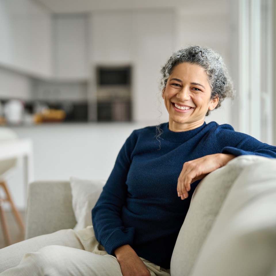Happy middle aged woman sitting on comfy sofa in living room at home laughing. Smiling mature older lady relaxing on couch looking at camera on couch in modern cozy house. Portrait.