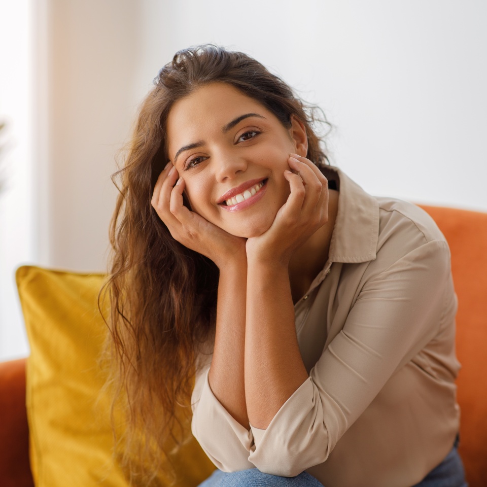 Beautiful young woman with wavy hair smiling and resting head on her hands while relaxing on comfortable couch in living room, happy female posing at camera while resting at home, free space