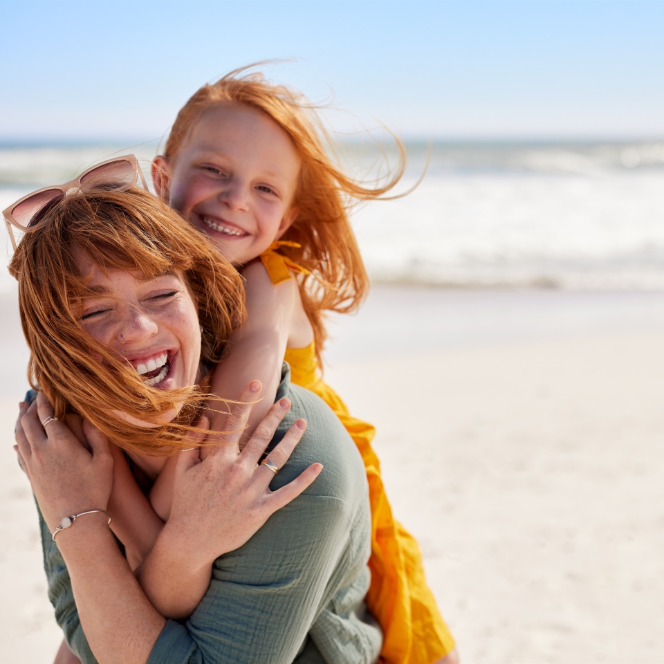 Smiling mother and beautiful daughter having fun at the beach. Portrait of happy woman giving a piggyback ride to cute little girl with copy space. Portrait of happy red hair kid embracing her mom.